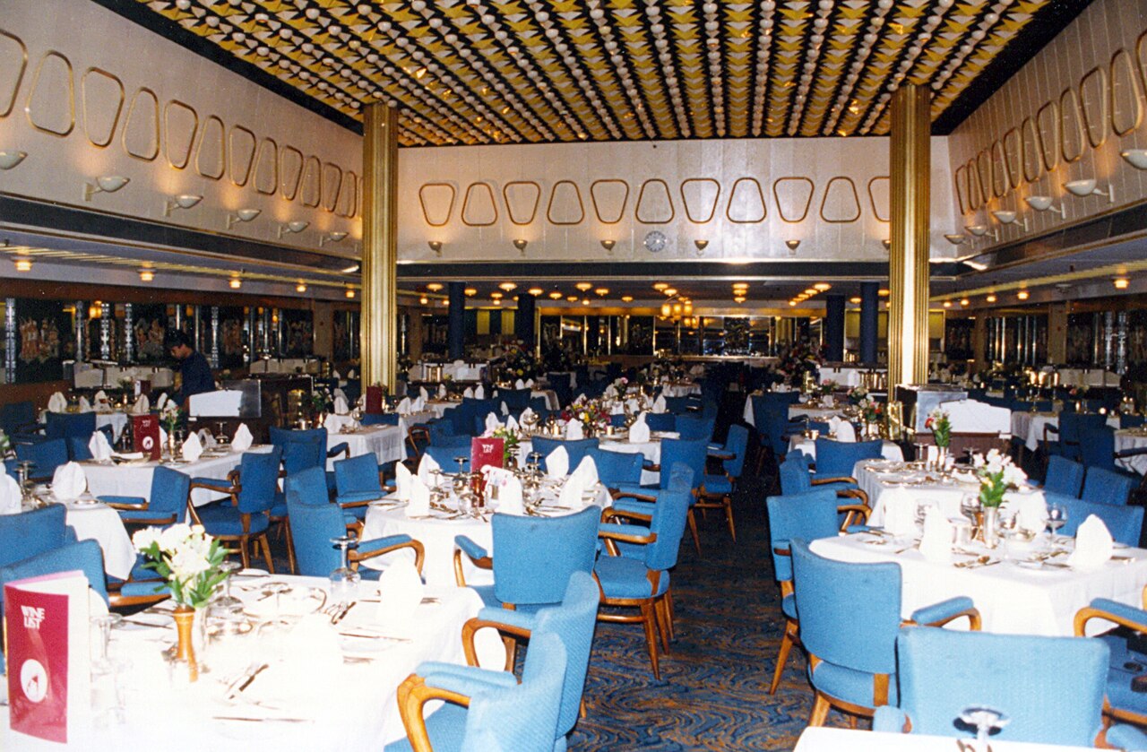 A ship's dining room with set tables and blue chairs.
