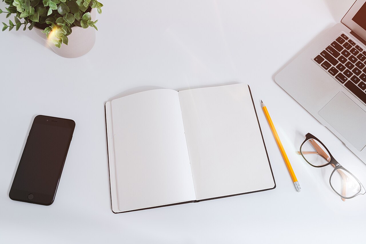 An open notebook, phone, glasses and laptop on a white desk.