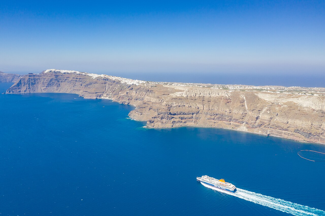 A ferry crossing the sea below the steep coast of a Greek island.