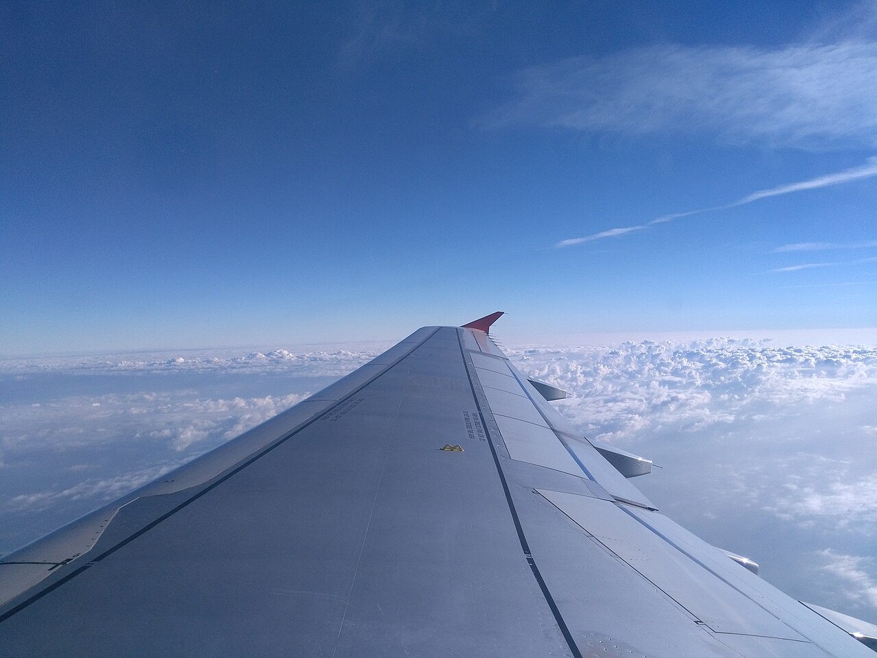 An aircraft wing above clouds beneath a blue sky.