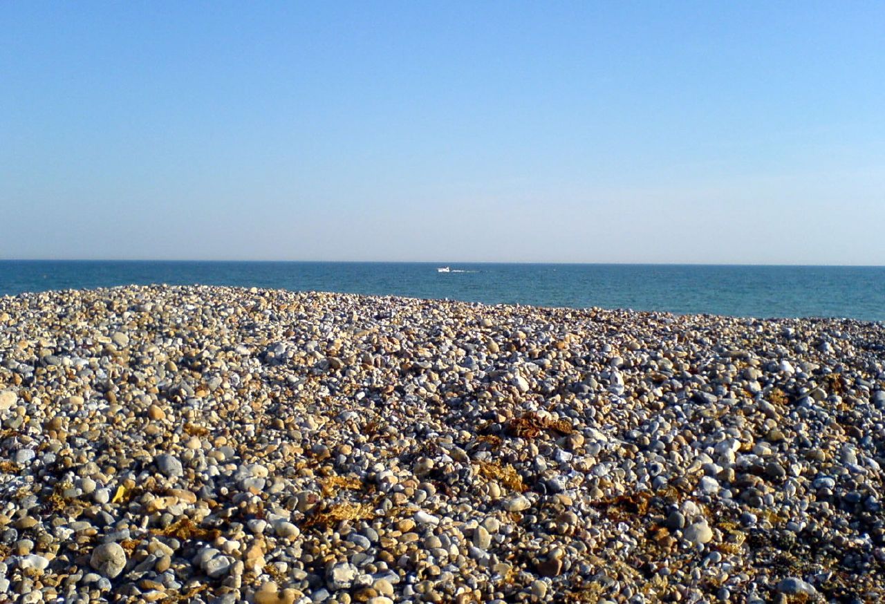 A pebble beach beneath a blue sky beside calm water.