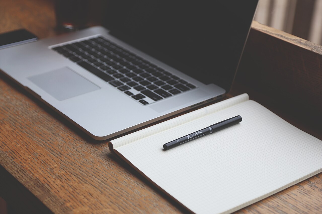 A laptop, blank notebook and pen on a wooden table.